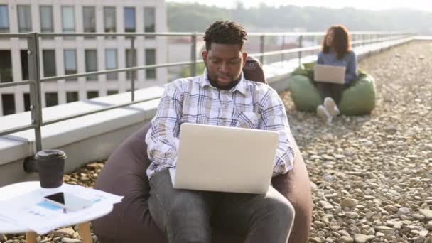 迷人的非洲裔美国男性自由职业者在休息时坐在布袋椅上 在办公室顶部用手提电脑进行远程工作 女商人同事的背景模糊不清 — 图库视频影像