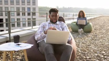 Binanın çatısındaki laptop kamerasına günlük kıyafet jesti yapan mutlu Afrikalı Amerikalı erkek, arka planda bulanık çalışan bayan meslektaşı....