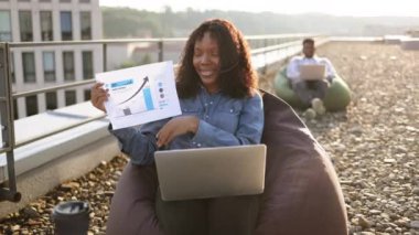 Panoramik terasta mali veri planını tartışan kot gömlekli ve kulaklı sakin Afrikalı Amerikalı iş kadını kameraya bakarken, erkek meslektaşım da bulanık arka plan üzerinde çalışıyor..