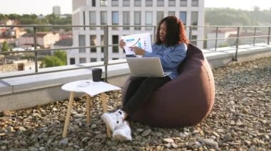 Günlük giysiler içinde genç Afrikalı bir kadın ofis dışında kulaklık ve bilgisayar kullanarak online sunum yapıyor. Terasta bilgi grafikleri tarafından gösterilen istatistikler hakkında uzaktan yönetici beyin fırtınası.