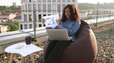 Günlük giysiler içinde genç Afrikalı bir kadın ofis dışında kulaklık ve bilgisayar kullanarak online sunum yapıyor. Terasta bilgi grafikleri tarafından gösterilen istatistikler hakkında uzaktan yönetici beyin fırtınası.