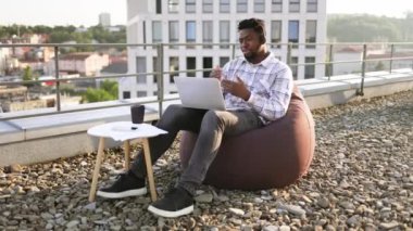 Gevşemiş Afrikalı Amerikalı adam, düz çatıda kalırken bilgisayar ve kulaklıkla internet konferansı düzenliyor. Şirket ofislerinin dışında kablosuz cihazlarla çalışan profesyonel girişimci..