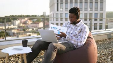 Günlük kıyafetler içindeki genç Afrikalı erkek ofis dışında kulaklık ve bilgisayar kullanarak online sunum yapıyor. Terasta bilgi grafikleri tarafından gösterilen istatistikler hakkında uzaktan yönetici beyin fırtınası.