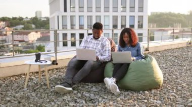 Ofisin çatısındaki puf minderlerde otururken dizüstü bilgisayar kullanan genç ve güzel bir Afro-Amerikan çift. Çağdaş cihazlarla çalışan iki iş adamı ofis dışındaki terasta oturuyor..