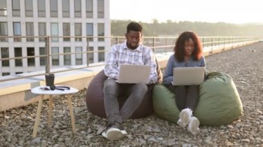 Ofisin çatısındaki puf minderlerde otururken dizüstü bilgisayar kullanan genç ve güzel bir Afro-Amerikan çift. Çağdaş cihazlarla çalışan iki iş adamı ofis dışındaki terasta oturuyor..