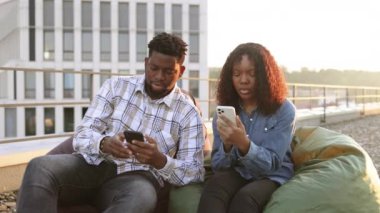 İki Afrikalı Amerikalı iş adamı, erkek ve kadın öğle yemeği molasında ofisin çatısındaki puf minderlerde oturuyor, cep telefonlarına bakıyor ve eğlenceli bilgiler paylaşıyorlar..