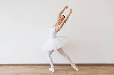 Beyaz etekli beyaz kadın balerin, kolları yukarıda dans ediyor, stüdyoda modern stüdyo salonunun parlak arka planına karşı..