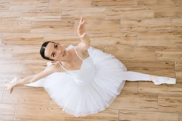 Top view of professional caucasian ballerina in white tutu and pointe stretching while raising hands over her head on wooden floor of studio hall. Copyspace.