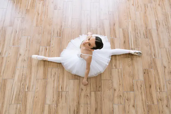 Gorgeous Caucasian ballerina sitting on the floor of bright studio. Young woman ballerina in white pack posing on background of wooden floor.
