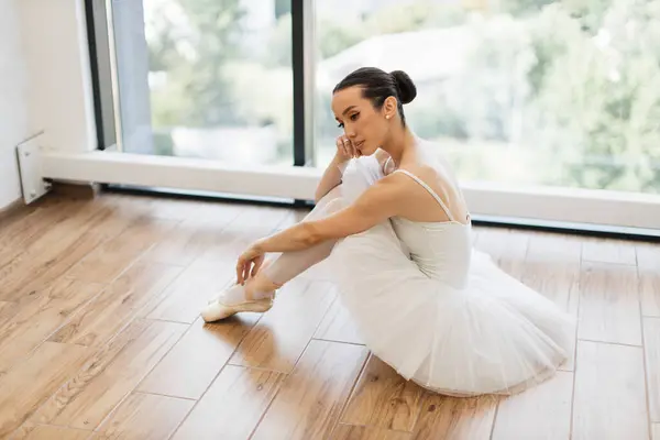Side view portrait of talented ballerina finished her wonderful performance and resting while sitting at wooden floor, copy space. Beautiful young female ballet dancer relaxing after rehearsal.
