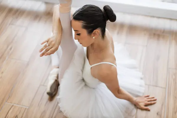 Ballet dancer wraps around the leg with hand and touching to head on background of panoramic window. Young and incredibly beautiful ballerina sitting at wooden studio floor and stretching.