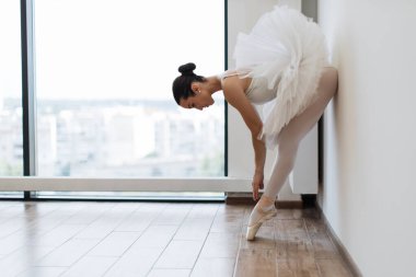 Beyaz balerin plie pozisyonunda zarif bir balerin ışık salonundaki geniş pencerenin yanında. Bale sınıfı eğitimi, kopyalama alanı. Klasik Kafkas Bale tarafı görünümü.