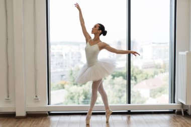 Dans sınıfındaki beyaz, klasik balerin genç bayan. Güzel, zarif balerin, bale pozisyonu beyaz etekli, beyaz ışıklı salonun geniş penceresinin yanında..