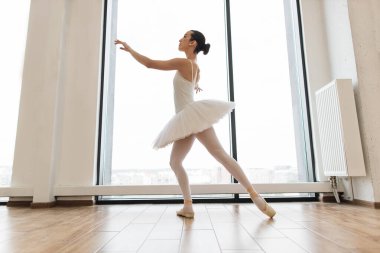 Beyaz salonda geniş pencerenin yanında beyaz etekli güzel balerin pozisyonları. Genç balerin - Bale stüdyosunda etekli uyumlu güzel kadın - Çağdaş dans sanatçısı.