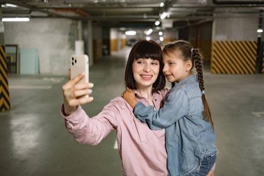 Mutlu bir aile, günlük kıyafetlerini giyip heyecan verici bir geziden önce cep telefonuyla görüntülü arama yapıyorlar. Beyaz bir anne, anaokulu kızıyla el ele tutuşup yeraltı otoparkında akıllı telefonuyla selfie çekiyor..
