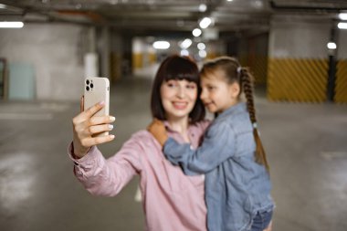 Mutlu bir aile, günlük kıyafetlerini giyip heyecan verici bir geziden önce cep telefonuyla görüntülü arama yapıyorlar. Beyaz bir anne, anaokulu kızıyla el ele tutuşup yeraltı otoparkında akıllı telefonuyla selfie çekiyor..