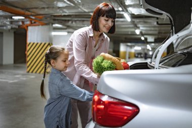 Çekici bir kadın ve küçük bir kız yeraltı otoparkında bagaja sebze dolu kağıt torba koyuyorlar. Kafkasyalı anne ve kızı süpermarketten alışverişlerini tamamlamışlar..