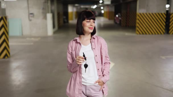 地下駐車場に立っている魅力的な白人女性の肖像画 遠隔操作で車を開けるキーを握るカメラを見るカジュアルな服 — ストック動画