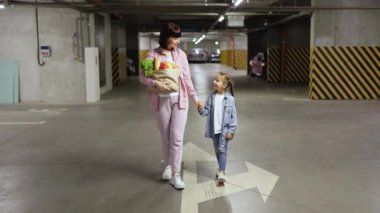 Modern yeraltı otoparkında ellerinde sebzelerle yürüyüp arabalarını arayan günlük kıyafetlerle gezen mutlu beyaz aile annesi ve kızı..