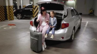 Kafkas anne ve küçük kızı yeraltı otoparkında modern bir arabanın bagajında oturuyorlar. Çocuklu genç bir kadın seyahat çantası taşıyor. Tatil planlamak için telefon kullanıyor..