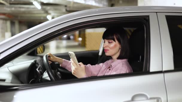 年轻自信的白种人女人穿着休闲装坐在现代汽车的轮子后面 手持智能手机 地下停车场灰色车辆内的电话中迷人的女性打字 — 图库视频影像