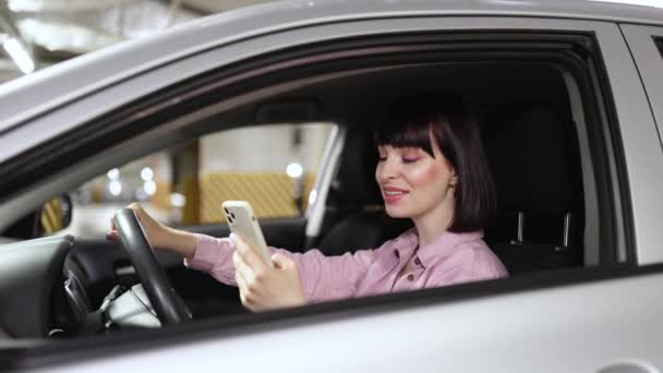 在地下停车场灰色车辆内的电话中 吸引人的女性打字 年轻自信的白种人女人 穿着休闲装 坐在现代汽车的轮子后面 手持智能手机 — 图库视频影像