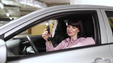 Kendine güveni tam beyaz bir kadın modern bir arabanın direksiyonunda oturuyor yeraltı otoparkında elinde anahtarı tutuyor ve kameraya bakıyor..