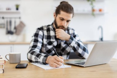 Verimli iş sahibi taşınabilir bilgisayardaki verileri kontrol ederken sözleşmeyi geliştiriyor. Gün ortasında evde otururken dokümana kareli gömlek giyen yakışıklı beyaz adam.