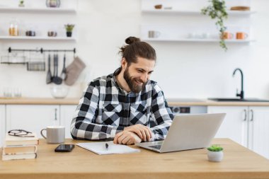 Düğmeli tişörtlü mutlu Kafkas profesyoneli evde otururken video görüşmesinde müşteriyle konuşuyor. Pozitif sakallı işletme yöneticisi internet uygulaması kullanarak projenin yeni detaylarını açıklıyor.