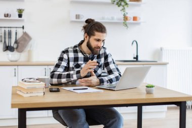 Ciddi bir serbest çalışan web konferansı sırasında etkili iletişim yeteneğini gösteriyor. Kablosuz kulaklık takan genç beyaz adam mutfak içerisindeki dizüstü bilgisayarla takım arkadaşlarıyla çevrimiçi toplantı yapıyor..