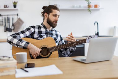 Geniş apartmandaki taşınabilir bilgisayar kamerasına gülümserken elinde telli enstrüman tutan neşeli beyaz insan. Modern teknolojileri kullanarak online gitar dersi veren mutlu bir erkek..