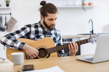 İnternet üzerinden telli çalgılar üzerinde ses egzersizleri yapmaya odaklanmış beyaz bir erkek. Dişli gömlekli genç müzisyen, akustik gitarını akustik dairede dizüstü bilgisayar kullanarak akort ediyor..