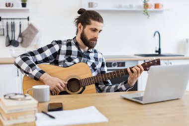 Geniş apartmandaki taşınabilir bilgisayar kamerasına gülümserken elinde telli enstrüman tutan neşeli beyaz insan. Modern teknolojileri kullanarak online gitar dersi veren mutlu bir erkek..