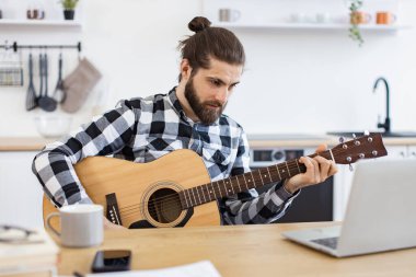 Modern teknolojileri kullanarak online gitar dersi veren mutlu bir erkek. Geniş apartmandaki taşınabilir bilgisayar kamerasına gülümserken elinde telli enstrüman tutan neşeli beyaz insan..