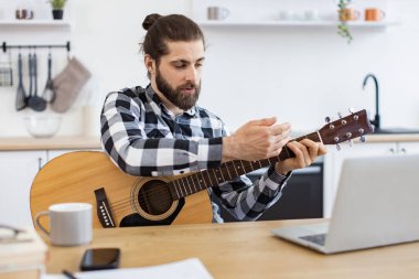 Modern teknolojileri kullanarak online gitar dersi veren mutlu bir erkek. Geniş apartmandaki taşınabilir bilgisayar kamerasına gülümserken elinde telli enstrüman tutan neşeli beyaz insan..