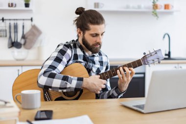Modern teknolojileri kullanarak online gitar dersi veren mutlu bir erkek. Geniş apartmandaki taşınabilir bilgisayar kamerasına gülümserken elinde telli enstrüman tutan neşeli beyaz insan..