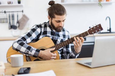 Evde eğlenirken altı telli gitarla poz veren yakışıklı sakallı yetişkin portresi. Gülümseyen beyaz adam bilgisayardan online dersler alarak enstrüman çalmayı öğreniyor..