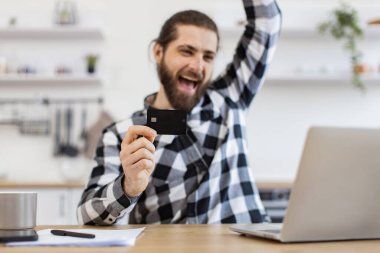 Duygusal sakallı yetişkin. Mutfaktaki bilgisayar ekranına bakarken heyecanlı bir şekilde elini kaldırıyor. Beyaz erkek internet bankacılığı kullanarak başarılı bir alışveriş yapıyor..