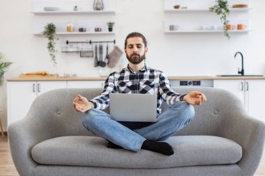 Oturma odasında taşınabilir dizüstü bilgisayar üzerinde çalışırken gözleri kapalı meditasyon yapan beyaz bir adam. Sakallı serbest çalışan, rahat koltukta oturan, yoga yaparak yorgunluğu azaltan biri..