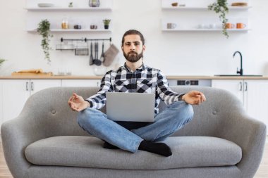 Oturma odasında taşınabilir dizüstü bilgisayar üzerinde çalışırken gözleri kapalı meditasyon yapan beyaz bir adam. Sakallı serbest çalışan, rahat koltukta oturan, yoga yaparak yorgunluğu azaltan biri..