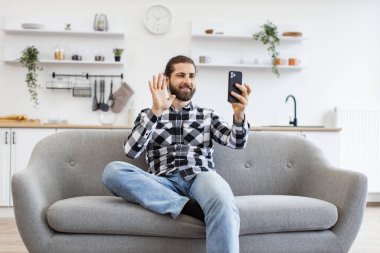 Kareli tişörtlü, sakallı bir yetişkin, mutfakta koltukta otururken göz hizasında akıllı telefon kaldırırken selam veriyor. Neşeli iş adamı, çalışma saatleri boyunca video çağrılarına katılanları selamlıyor