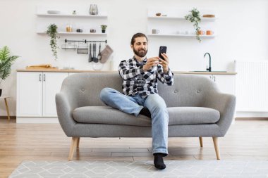 Mutfakta web kamerasıyla konuşurken telefonu tutan çekici bir genç adam. Damalı gömlekli mutlu Kafkasyalı ev içlerindeki koltukta oturan modern bir cihazla online konferans düzenliyor..