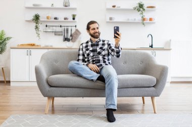 Mutfakta web kamerasıyla konuşurken telefonu tutan çekici bir genç adam. Damalı gömlekli mutlu Kafkasyalı ev içlerindeki koltukta oturan modern bir cihazla online konferans düzenliyor..