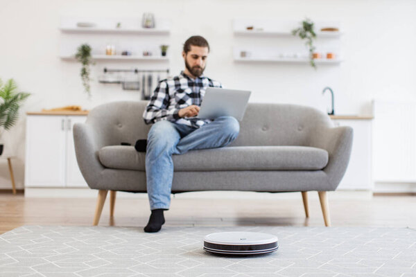 Focus on smart robotic vacuum operated by happy Caucasian man while performing automatic cleaning on carpet indoors. Cheerful owner programming home gadget via remote control while focusing on work.