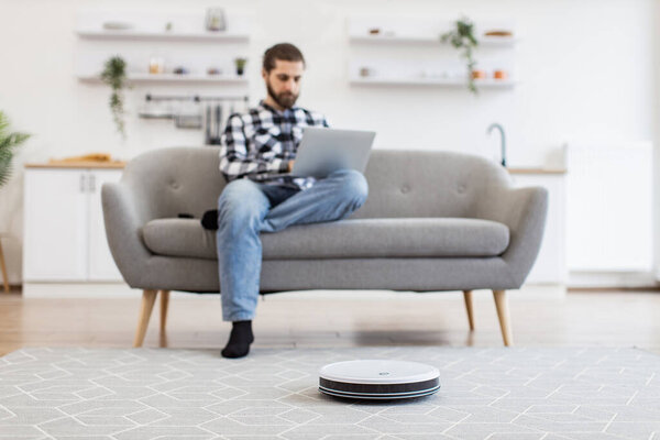 Robotic vacuum removing dust from carpet while Caucasian homeowner utilizing laptop sitting on sofa. Relaxed freelancer leading comfortable lifestyle by using tech gadgets for home kitchen.