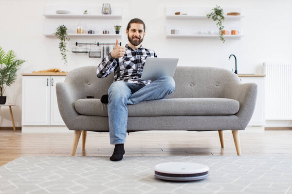 Relaxed freelancer leading comfortable lifestyle by using tech gadgets for home kitchen. Robotic vacuum removing dust from carpet while Caucasian homeowner utilizing laptop sitting on sofa.