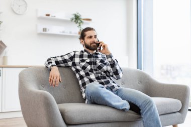 Geniş yemek odasındaki kanepede otururken akıllı telefondan konuşan sıradan giyinmiş neşeli genç adam. Uzaktan kumandalı bir şirketin müşterisiyle verimli bir iş görüşmesi yapan Kafkasyalı serbest çalışan..