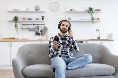 Evde dijital cihazlar üzerinden müzik dinlemeden önce sakin ev sahibi çalma listesini kontrol ediyor. Kafkasyalı rahat bir insan, gündelik giysiler ve kulaklıklar takıyor. Elinde akıllı telefon, mutfakta koltukta oturuyor..
