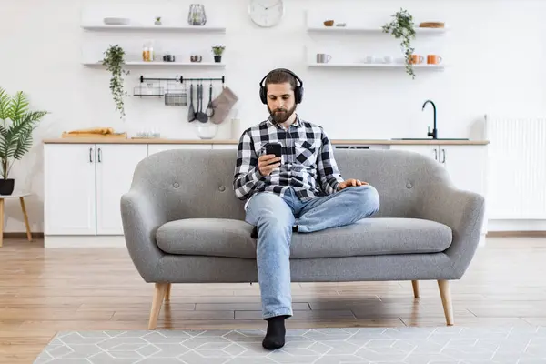 Relaxed Caucasian person in casual wear and headphones holding smartphone while taking seat at sofa in kitchen. Calm homeowner checking playlist before listening to music over digital devices at home.