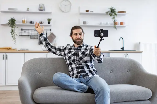 Happy Caucasian person conducting online conference via modern device sitting on couch in home interior. Attractive young man holding phone and selfie stick while talking via webcam on kitchen.